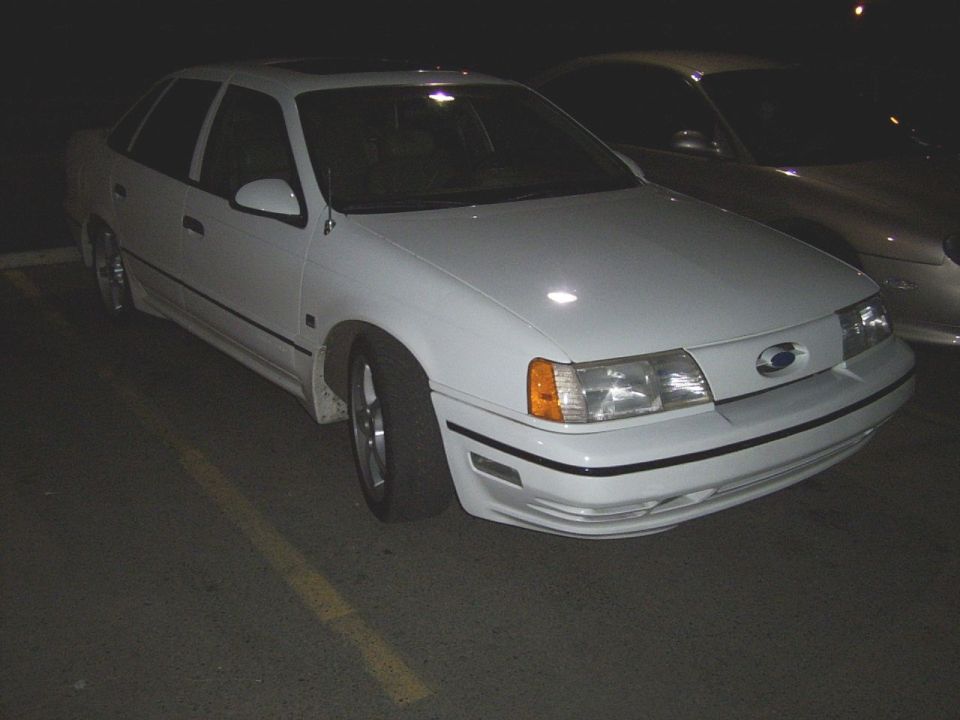 White sedan parked at night, another car behind it. Looks like a 1992 Ford Taurus