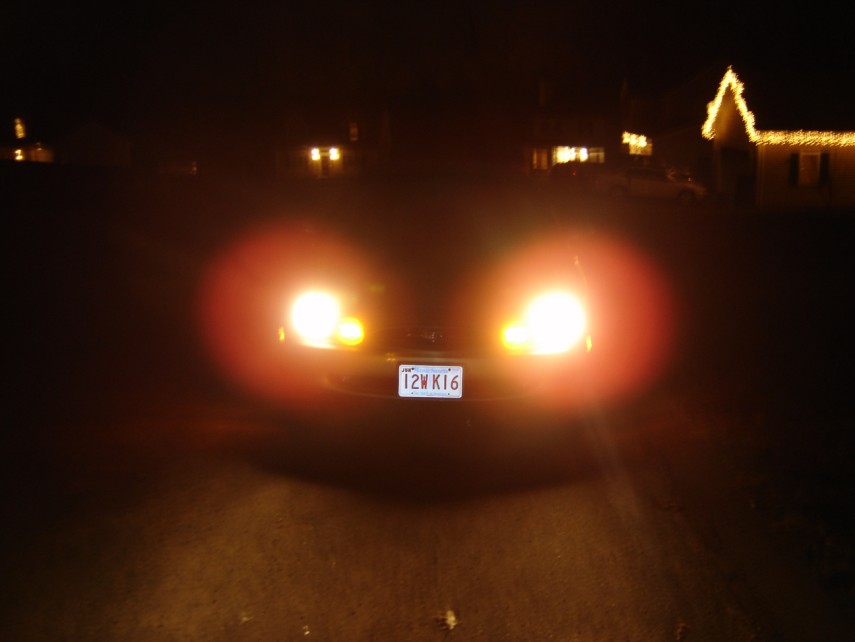 Front of car at night with headlights glowing, Massachusetts plate 12WK16, holiday lights behind
