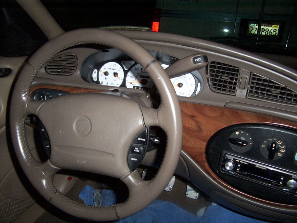 Car interior showing steering wheel, dashboard with wood trim, and radio. License plate visible in background