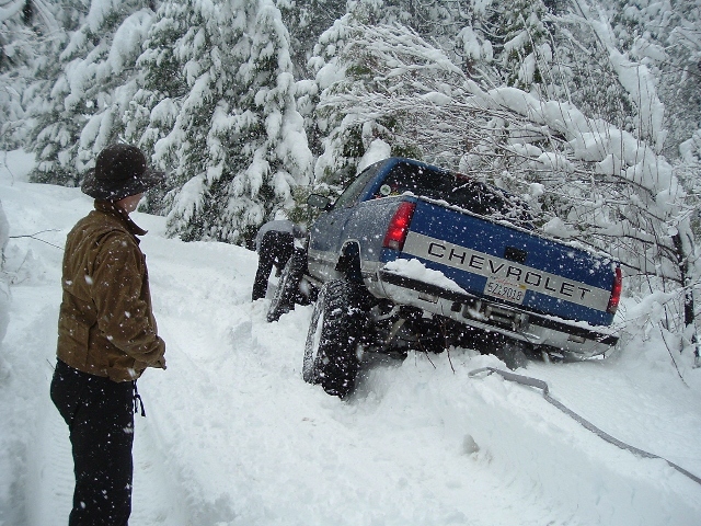 Lifted blue-and-white Chevrolet pickup stuck nose-down in deep snow as a person looks on