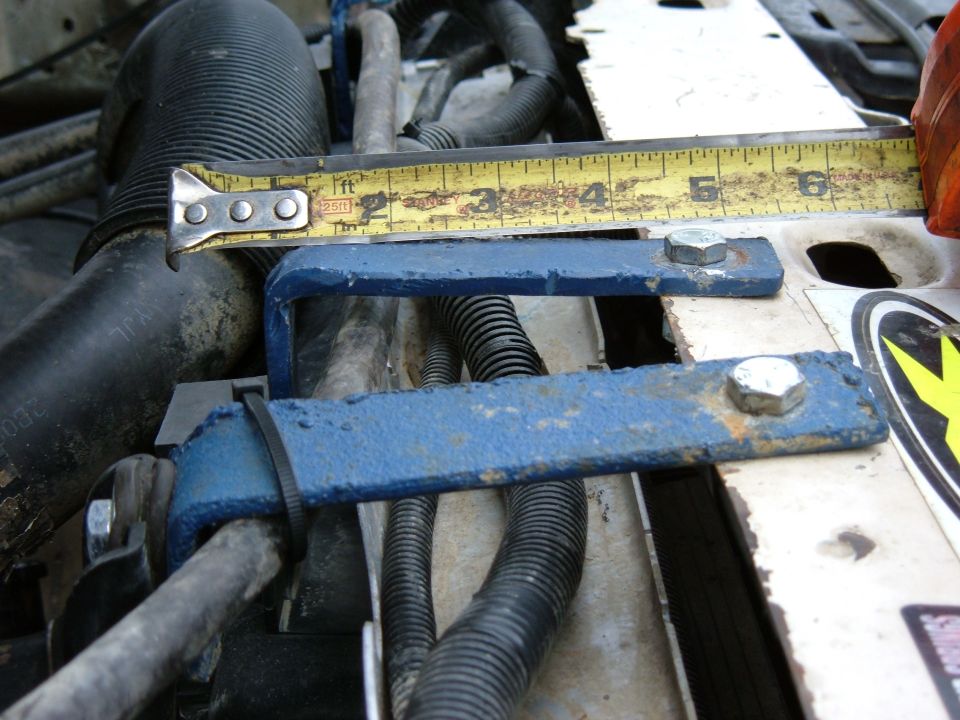 Engine bay with blue metal brackets and a tape measure. Black hoses and wiring are visible