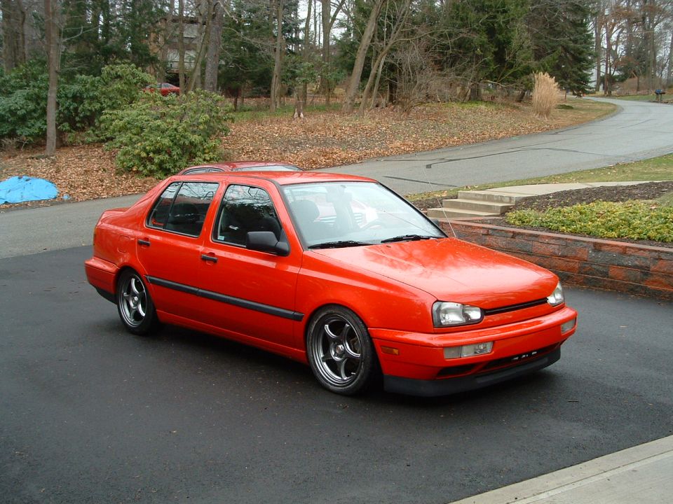 Red lowered VW Jetta Mk3 sedan on polished wheels, 3/4 front view on asphalt driveway