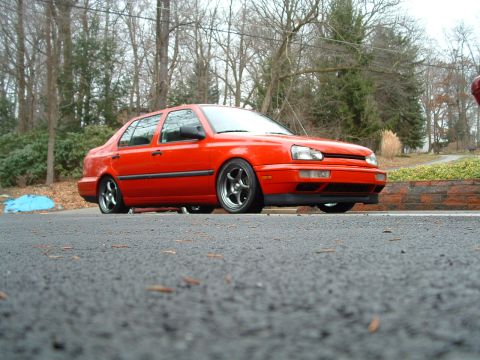 1994 Volkswagen Jetta - A low-slung orange sedan sits parked on a paved surface, with trees in the background.