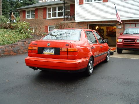 1994 Volkswagen Jetta - A red Volkswagen sedan is parked in front of a brick house with a red pickup truck visible in the garage.