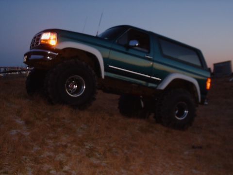 1996 Ford Bronco - A green 1996 Ford Bronco is shown in a 3/4 front view, driving on a grassy incline at dusk.