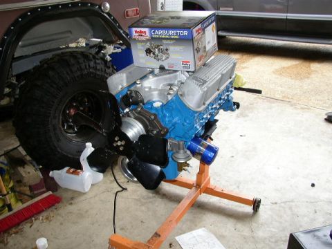 1995 Ford Bronco - A blue engine with a Holley carburetor box sits on an engine stand, with parts of two vehicles in the background.