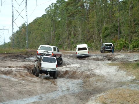 1995 Ford Bronco - Multiple SUVs and trucks tackle a challenging dirt trail with steep inclines and muddy patches.
