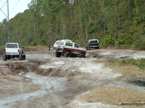 1995 Ford Bronco - Several off-road vehicles navigate a challenging dirt track in a wooded area.