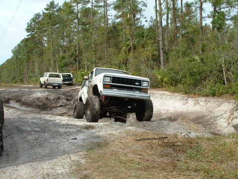 1995 Ford Bronco - A white SUV with significant suspension modifications tackles a challenging off-road course with other vehicles in the background.