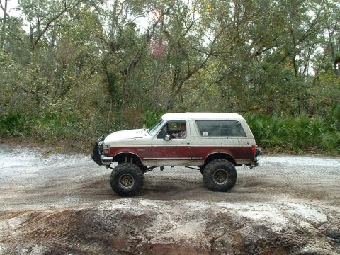 1995 Ford Bronco - This lifted 1995 Ford Bronco is shown off-roading on a dirt trail with trees in the background.