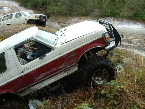 1995 Ford Bronco - Two Ford Broncos navigate a muddy off-road trail, with one vehicle partially submerged in the mire.