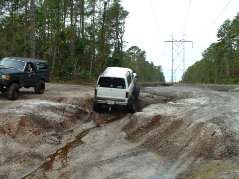 1995 Ford Bronco - Two Ford Broncos tackle a challenging off-road trail with muddy terrain and large ruts.