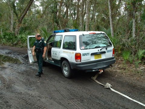 1995 Ford Bronco - Sheriff's Explorer SUV stuck in mud on a dirt road, deputy standing by. A tow strap is attached to the rear.