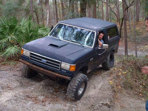 1995 Ford Bronco - A man drives a modified Ford Bronco through a wooded area with sandy terrain.