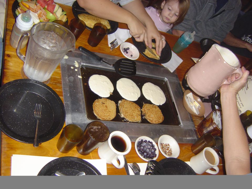 Family gathered around a table making pancakes on a griddle. A child watches as someone flips a pancake
