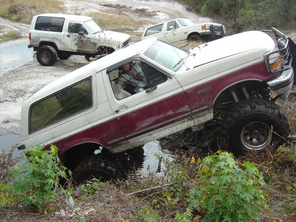 White and maroon 1993 Ford Bronco stuck in mud, front right wheel deep in water