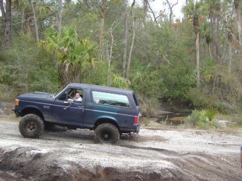1993 Ford Bronco - A 1993 Ford Bronco navigates a dirt trail, kicking up dust as it drives through a wooded area.