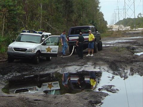 1993 Ford Bronco - Two vehicles, a police SUV and a lifted Bronco, are stuck in a muddy area with power lines in the background.