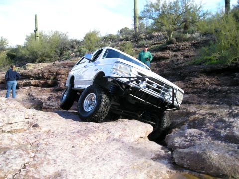 1993 Ford Bronco - A modified white Ford Bronco tackles a challenging rocky trail with two people observing.
