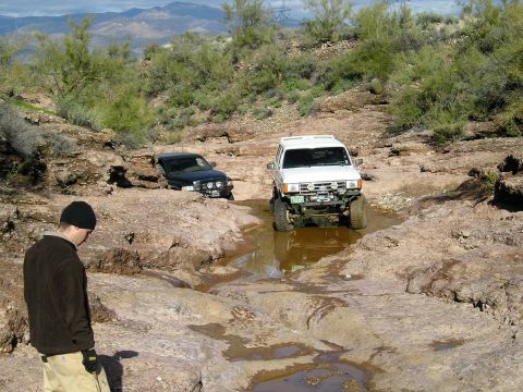 1993 Ford Bronco - Two SUVs navigate a challenging off-road course, with one vehicle partially submerged in mud.