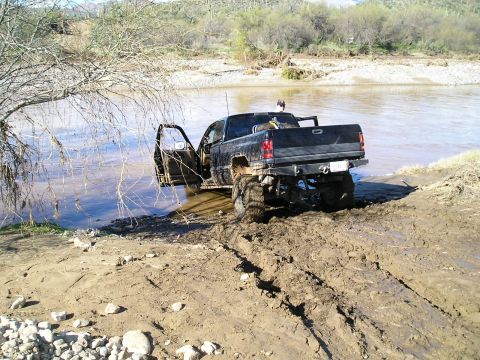 1993 Ford Bronco - A black pickup truck is stuck in the mud and water with its driver's door open, suggesting a difficult off-road situation.