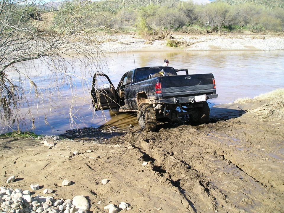 Mud-covered dark Dodge Ram pickup backing out of a river crossing with door open