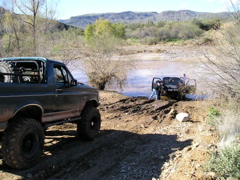 1993 Ford Bronco - Two off-road vehicles navigate a muddy river crossing in a desert landscape.