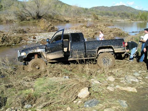 1993 Ford Bronco - A black pickup truck is stuck in a muddy riverbed, with people attempting to free it.