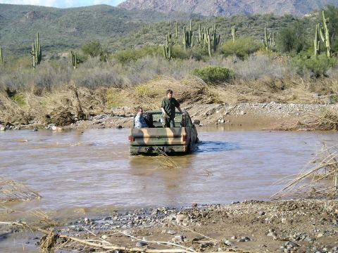 1993 Ford Bronco - A camouflaged pickup truck navigates a shallow river in a desert landscape, with two passengers in the truck bed.