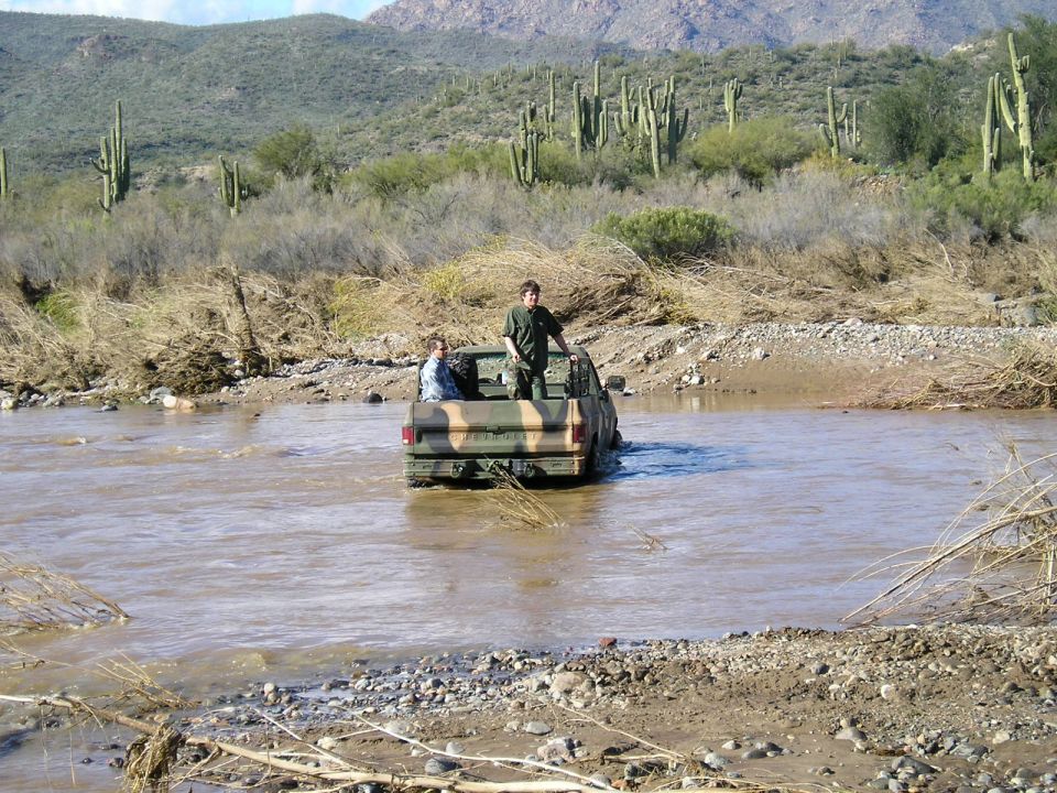 Camouflaged pickup truck driving through a river with two people in the bed