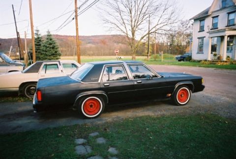 1989 Ford Crown Victoria LTD - A black sedan with red wheels sits parked on a gravel driveway next to another vehicle.