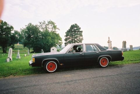 1989 Ford Crown Victoria LTD - A dark sedan with red wheels sits parked on the side of a road, adjacent to a cemetery.