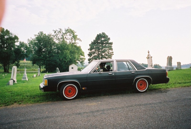Dark blue Ford Crown Victoria sedan with red wheels parked by a cemetery