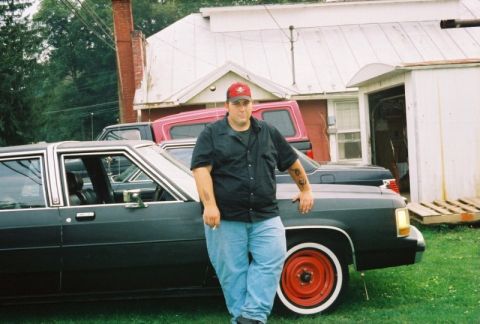 1989 Ford Crown Victoria LTD - A man stands next to a dark sedan with red wheels and white wall tires, with other vehicles and a building in the background.