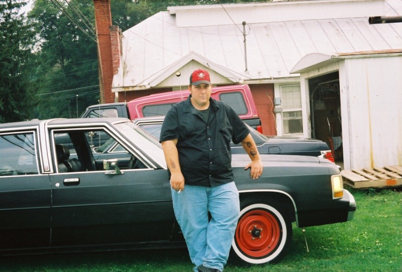 Man leaning on dark sedan with red steel wheel and whitewall tire on grass