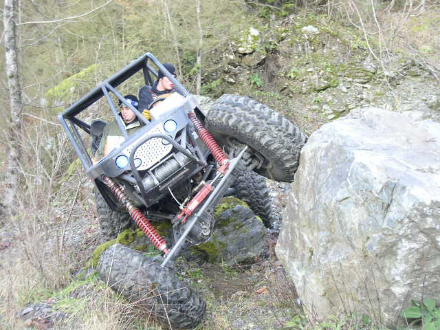 Grey tube-frame rock buggy with red coilovers crawling over mossy boulders