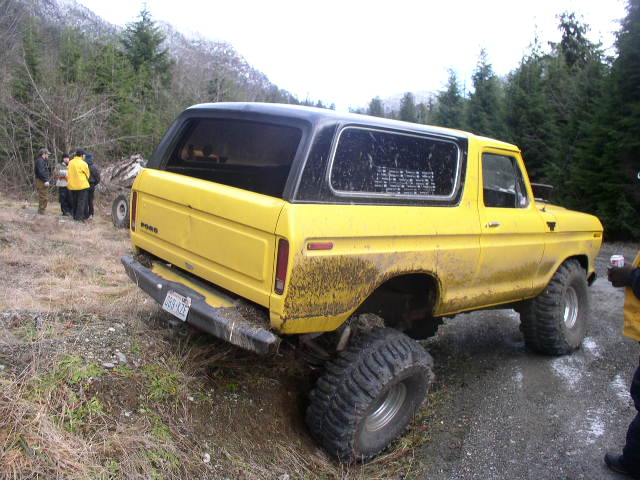 Muddy yellow lifted Ford Bronco with black top, rear 3/4 view on a gravel trail