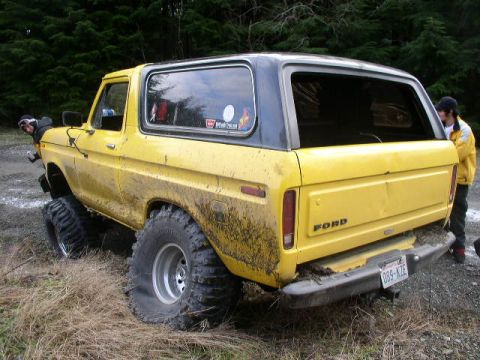1978 Ford Bronco - Three-quarter rear view of a mud-splattered yellow Ford Bronco with a black removable hardtop, FORD lettering across the tailgate and stick…