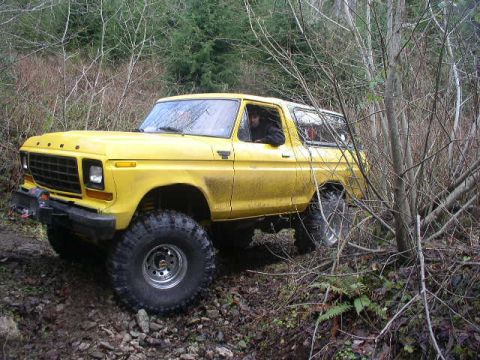1978 Ford Bronco - Driver-side 3/4 front view of a yellow lifted full-size Ford Bronco with the top removed, picking its way over rocks and roots on a wooded…