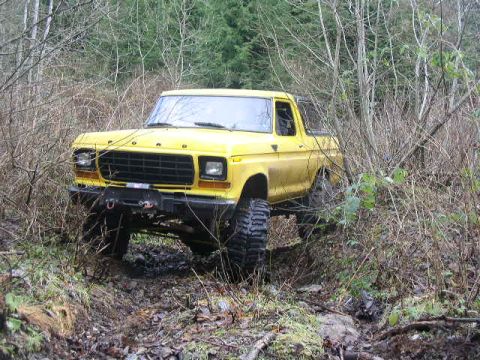 1978 Ford Bronco - Yellow lifted full-size Ford Bronco with the top removed, photographed from a low 3/4 front angle as it straddles a muddy rut on a wooded t…