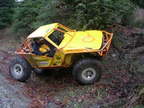 1978 Ford Bronco - A yellow tube-chassis rock buggy with a full orange exterior roll cage climbs off a gravel road onto a leaf-covered bank in the woods, driv…