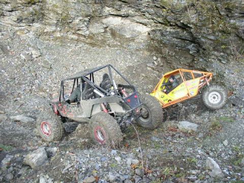 1978 Ford Bronco - Two tube-chassis rock buggies work up a loose rock face below a cliff: a tan and black open buggy with red coilovers and red-centered wheel…
