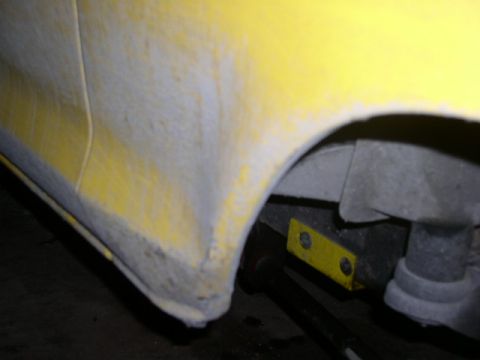 1978 Ford Bronco - Close-up of a yellow rear quarter panel and wheel arch stripped and sanded to gray primer along the lower lip, with the wheel removed. Behi…