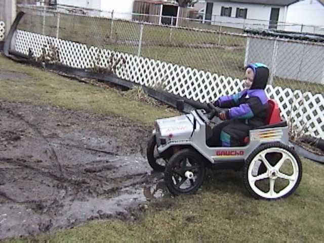 Child wearing a hooded jacket drives a silver toy jeep with 'GAUCHO' on the side through a muddy yard