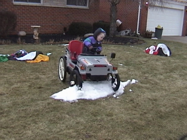 Toddler in winter coat sitting in gray and red Power Wheels ride-on jeep on a lawn over a patch of melting snow
