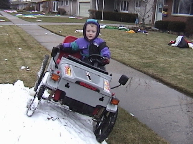 Child drives a toy jeep with 'BRONCO' written on the front, on a snowy sidewalk