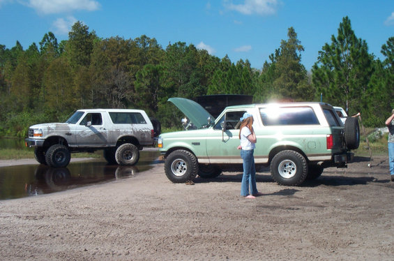 Mud-covered white Bronco beside a mint-green lifted Bronco with hood up on sandy lot
