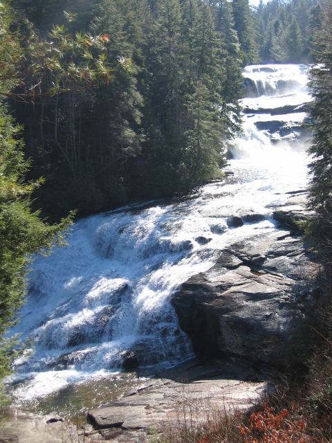 1985 Ford Crown Victoria LTD - A wide waterfall flows over rocky terrain, bordered by dense evergreen trees under a bright sky.