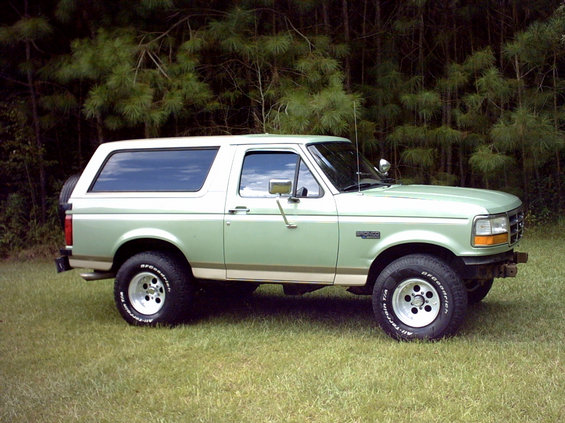 Green two-tone full-size Ford Bronco with white roof and chrome bumper parked on grass in front of pine trees