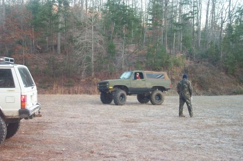 1987 Ford Bronco - A lifted, olive green SUV with large tires is parked in a dirt lot, with a white SUV and a person in camouflage nearby.